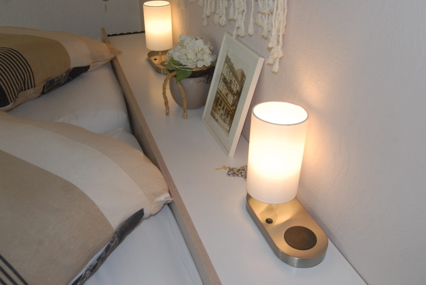 Handyladestation Ein Nachttisch mit zwei Lampen, einem Blumenarrangement und einem Bilderrahmen.