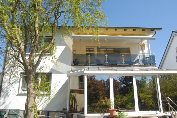 Helles modernes Haus mit Balkon und großer Fensterfront, umgeben von Bäumen.