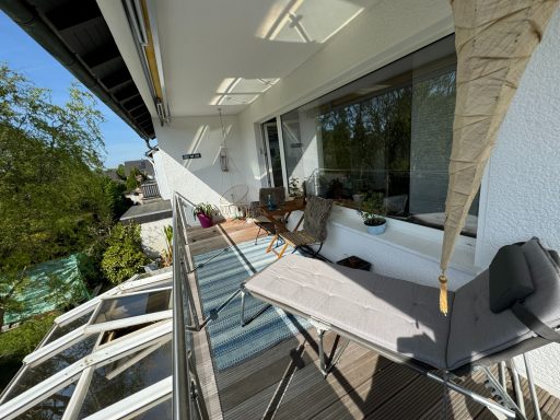 Heller Balkon mit Tisch, Stühlen und Sonnenschirm, umgeben von Bäumen und Grün.